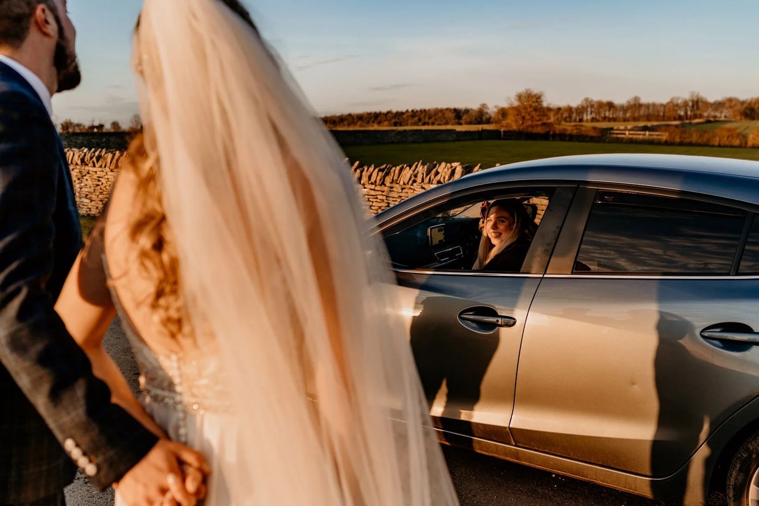 Wedding couple standing near luxury car during golden hour sunset on rural road.