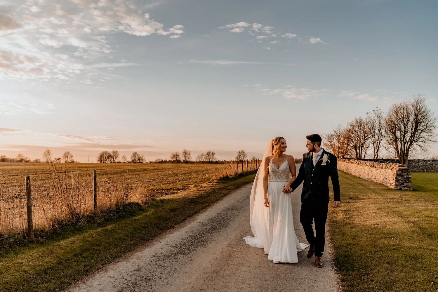 Silhouetted couple walks hand in hand through a golden prairie field at sunset with wispy clouds overhead.