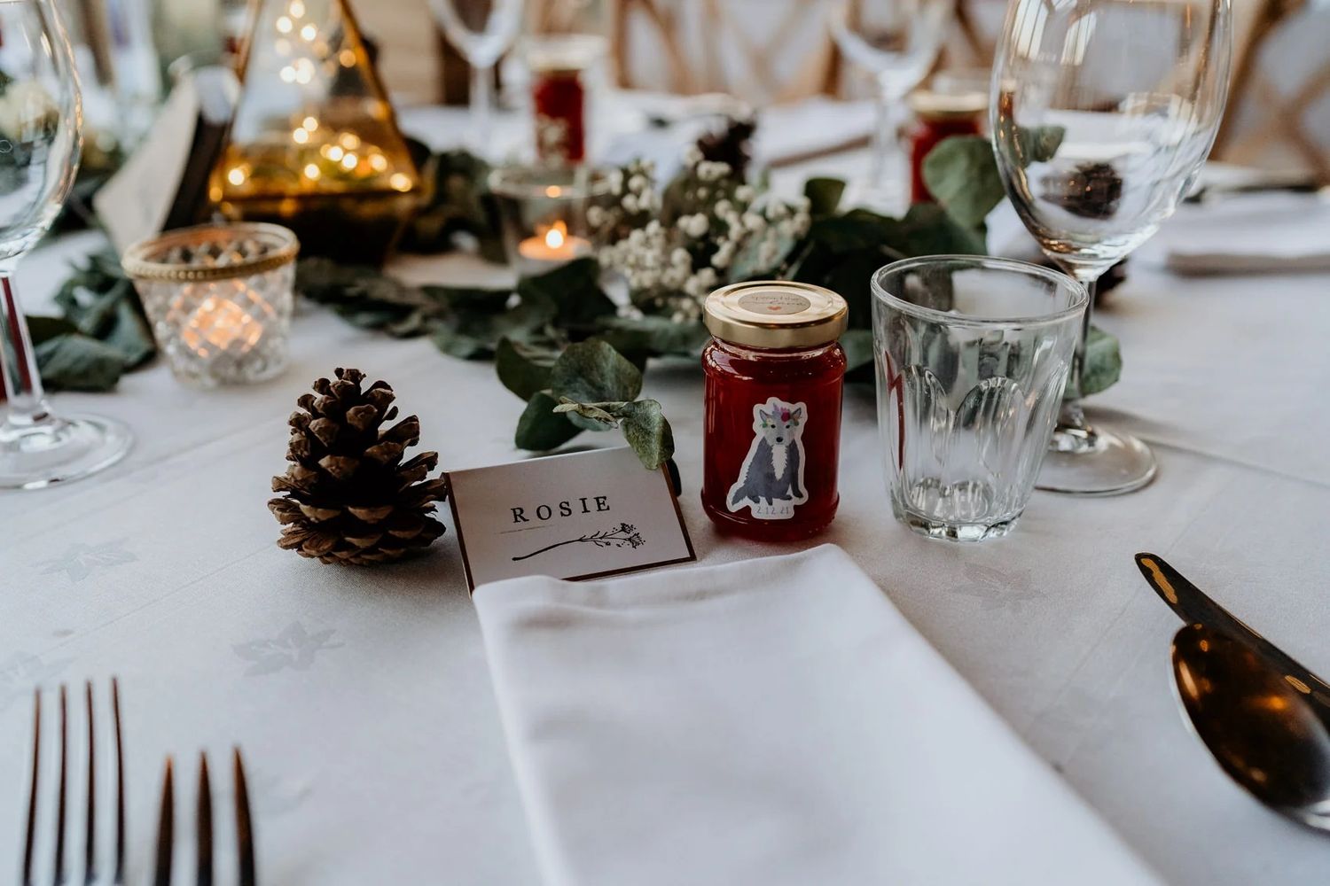 Wedding reception table decorated with pine cones, greenery, candles and place settings on white tablecloth.