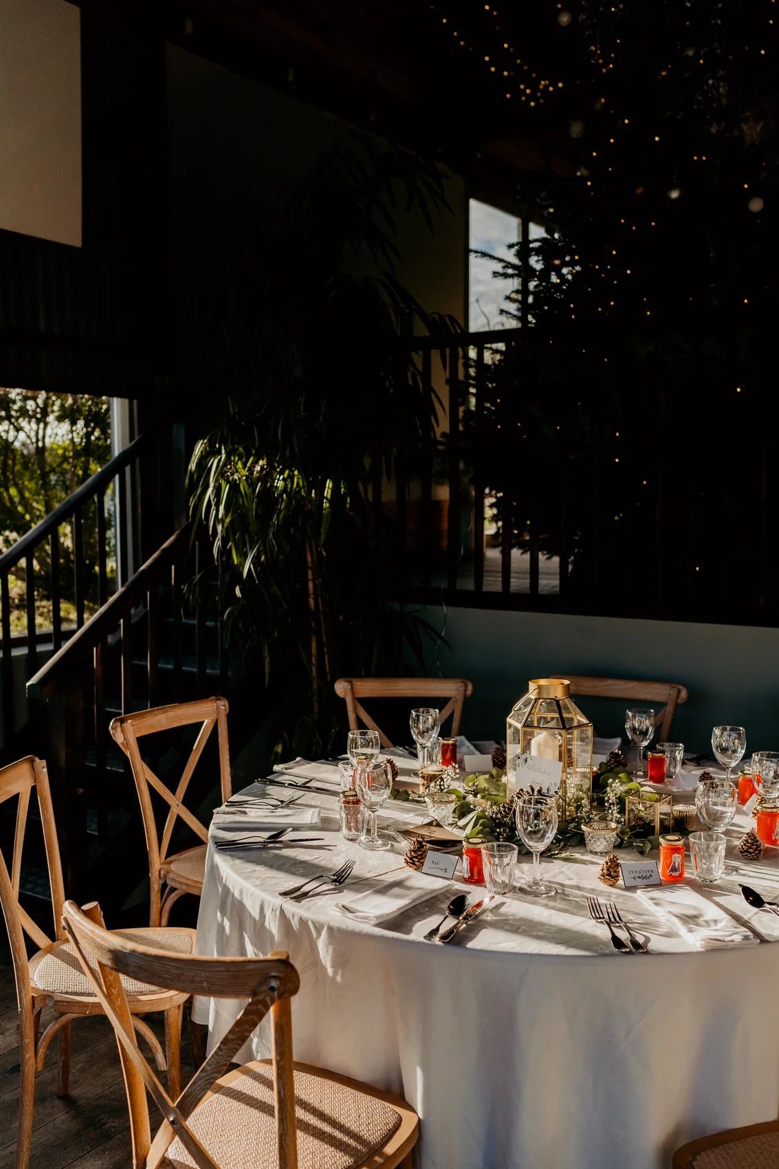 Elegant wedding reception tables set with white linens, wooden cross-back chairs, and ambient lighting in a cozy venue.