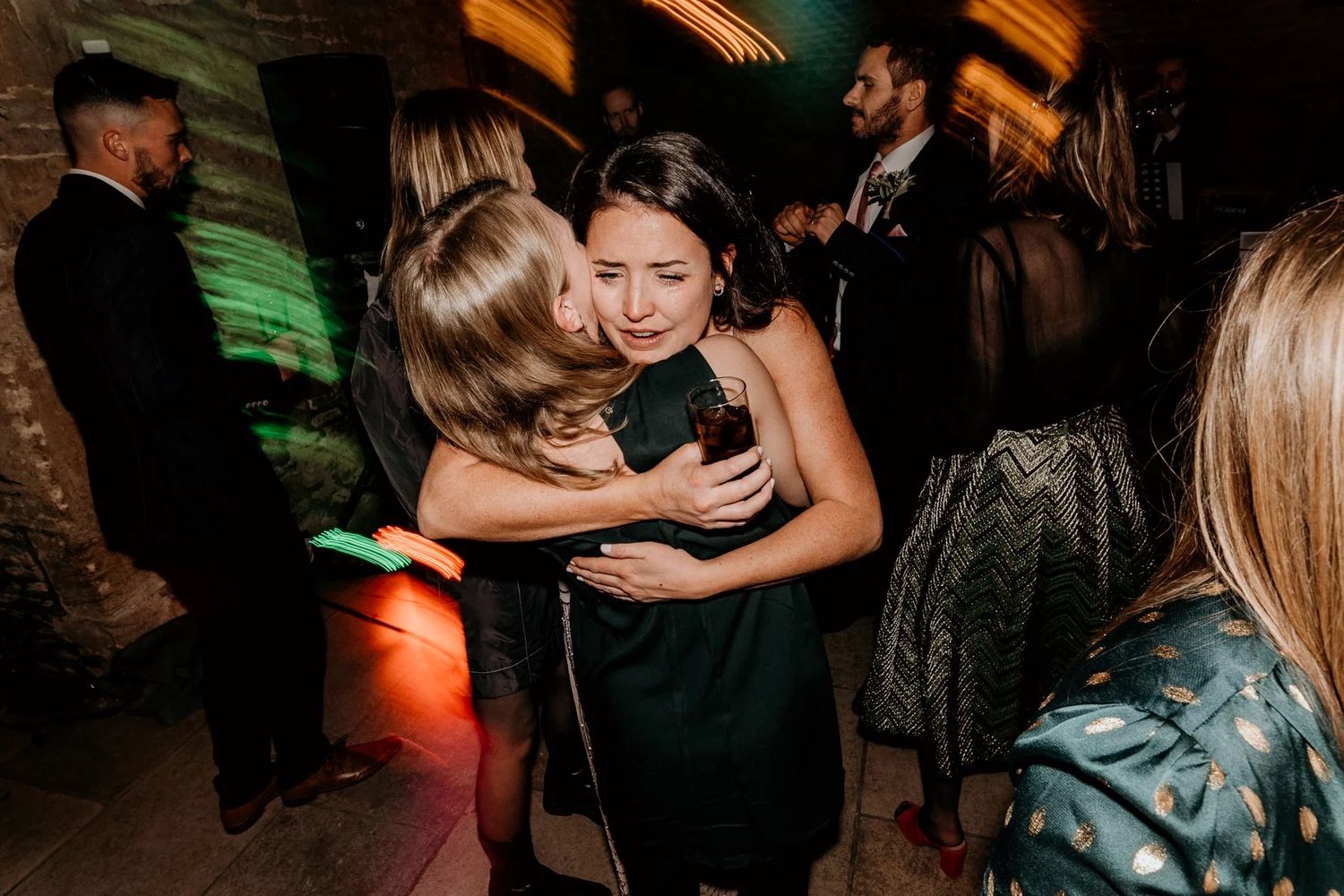 Group of wedding guests dancing together at a darkened reception venue with colorful lighting effects.