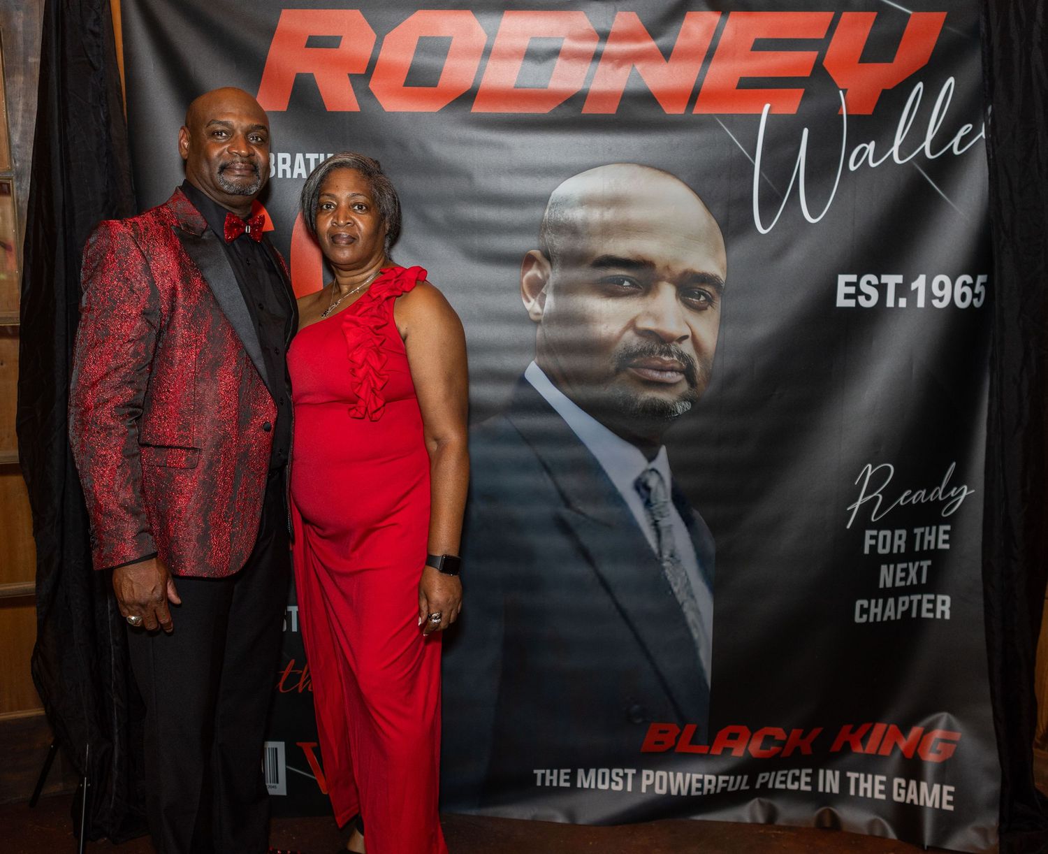 Formal occasion with red backdrop featuring couple in elegant attire posing in front of promotional banner.