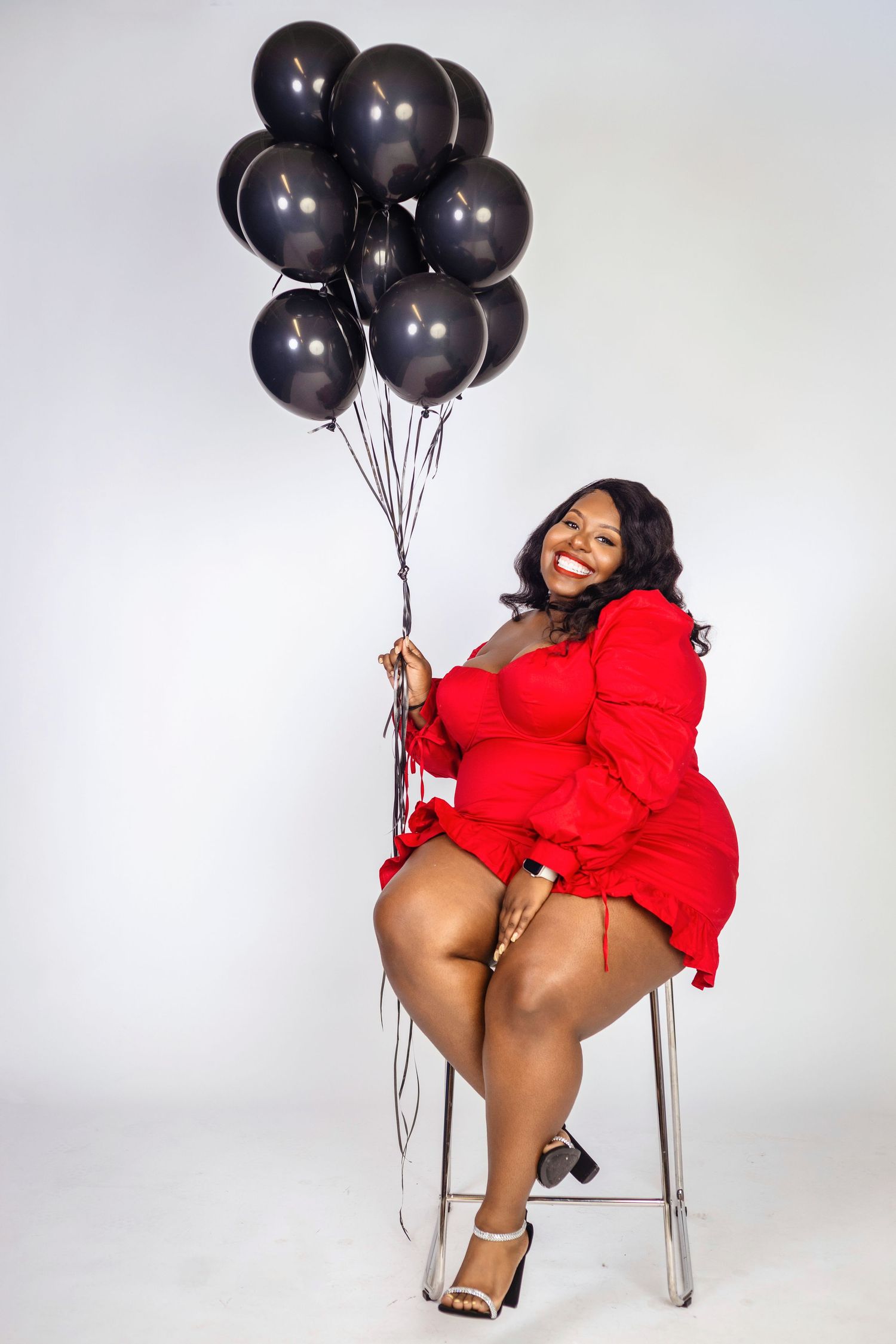 Stylish figure in red dress poses on stool with black balloons against white background.