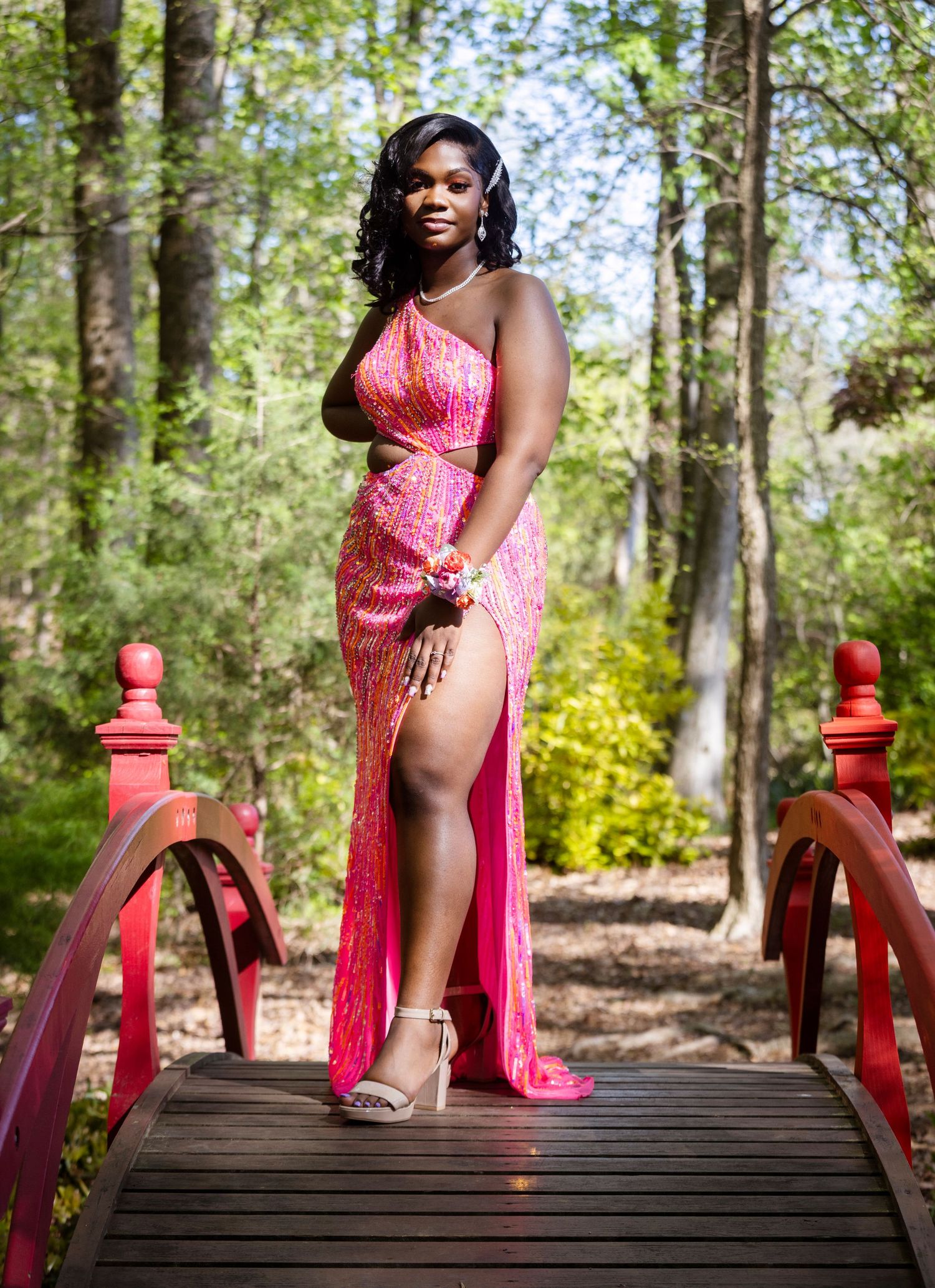 Woman in sparkling pink gown poses on red bridge in forest setting.