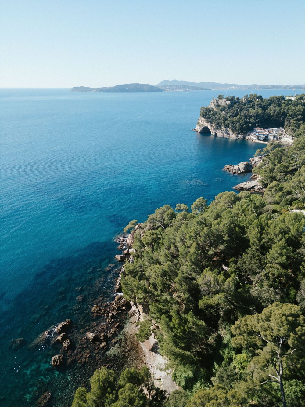 Aerial view of a rocky Mediterranean coastline with lush green trees, crystal blue sea, and distant islands under clear sky.