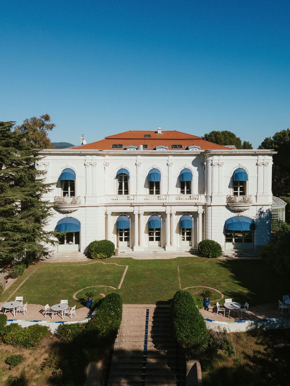 Aerial view of an elegant white neoclassical villa with terracotta roof, blue awnings, and manicured gardens under a clear blue sky.