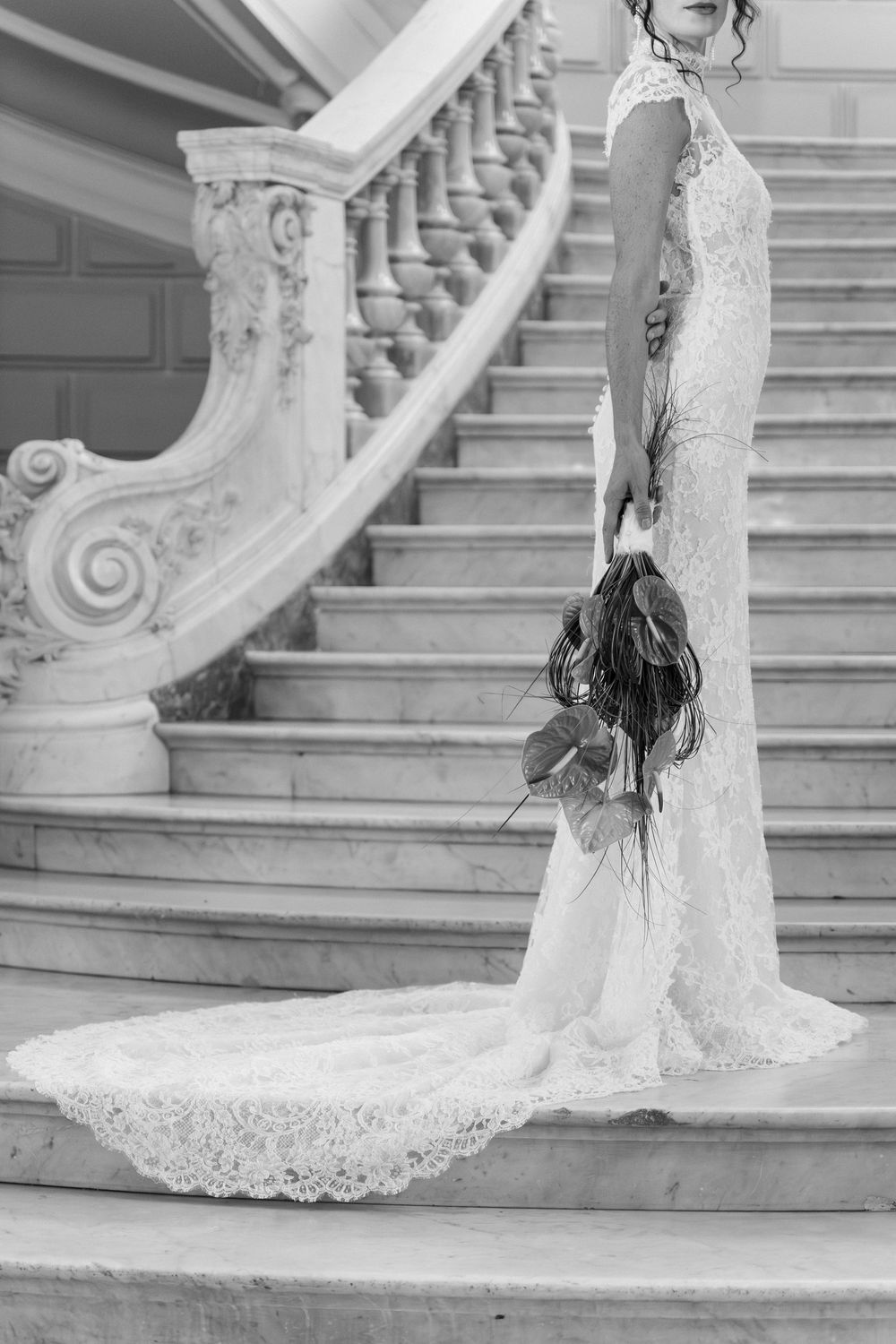 Bride in lace wedding gown with train holds bouquet on elegant marble staircase, black and white photo.