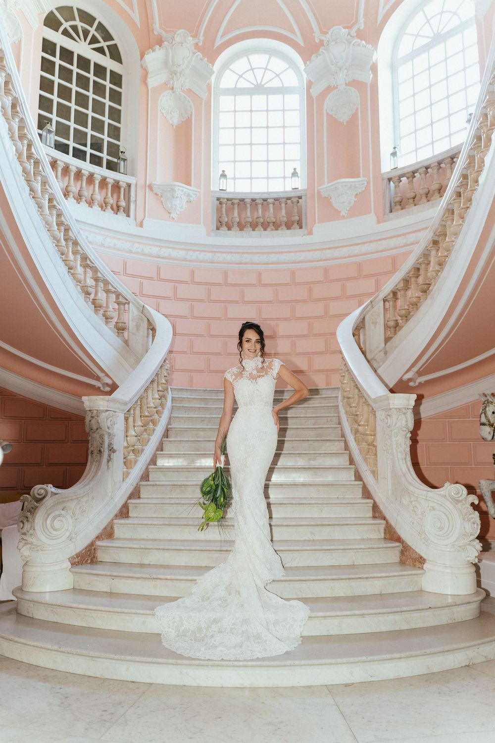 Bride in white lace mermaid gown holding bouquet on grand curved marble staircase in elegant pink ballroom.
