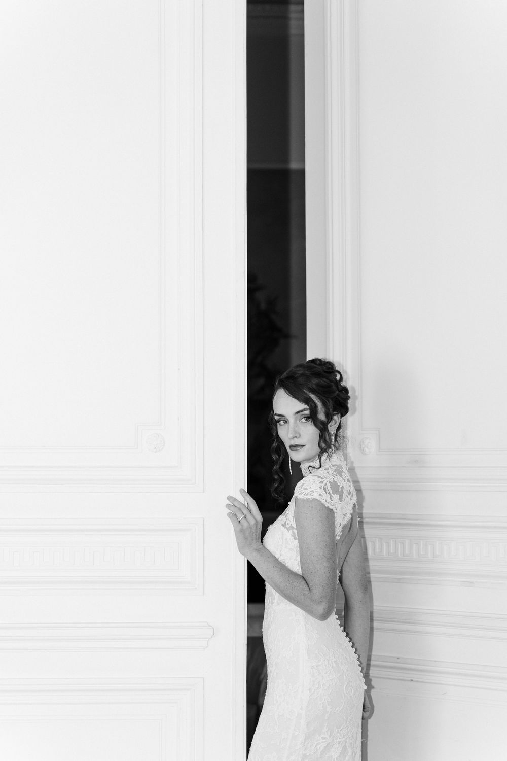 Elegant bride in lace wedding dress poses in doorway, black and white portrait photography.