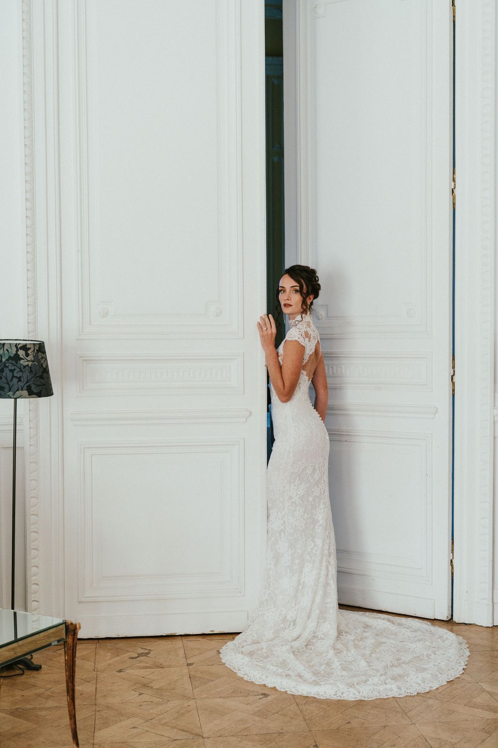 Bride in elegant lace wedding gown with train standing by white ornate doors in a classic interior.