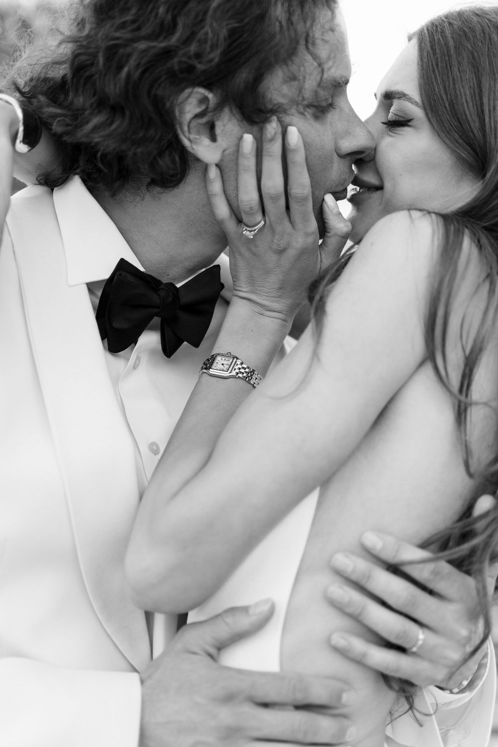Black and white photo of a couple sharing a romantic kiss, groom in white tuxedo with bow tie embracing bride.
