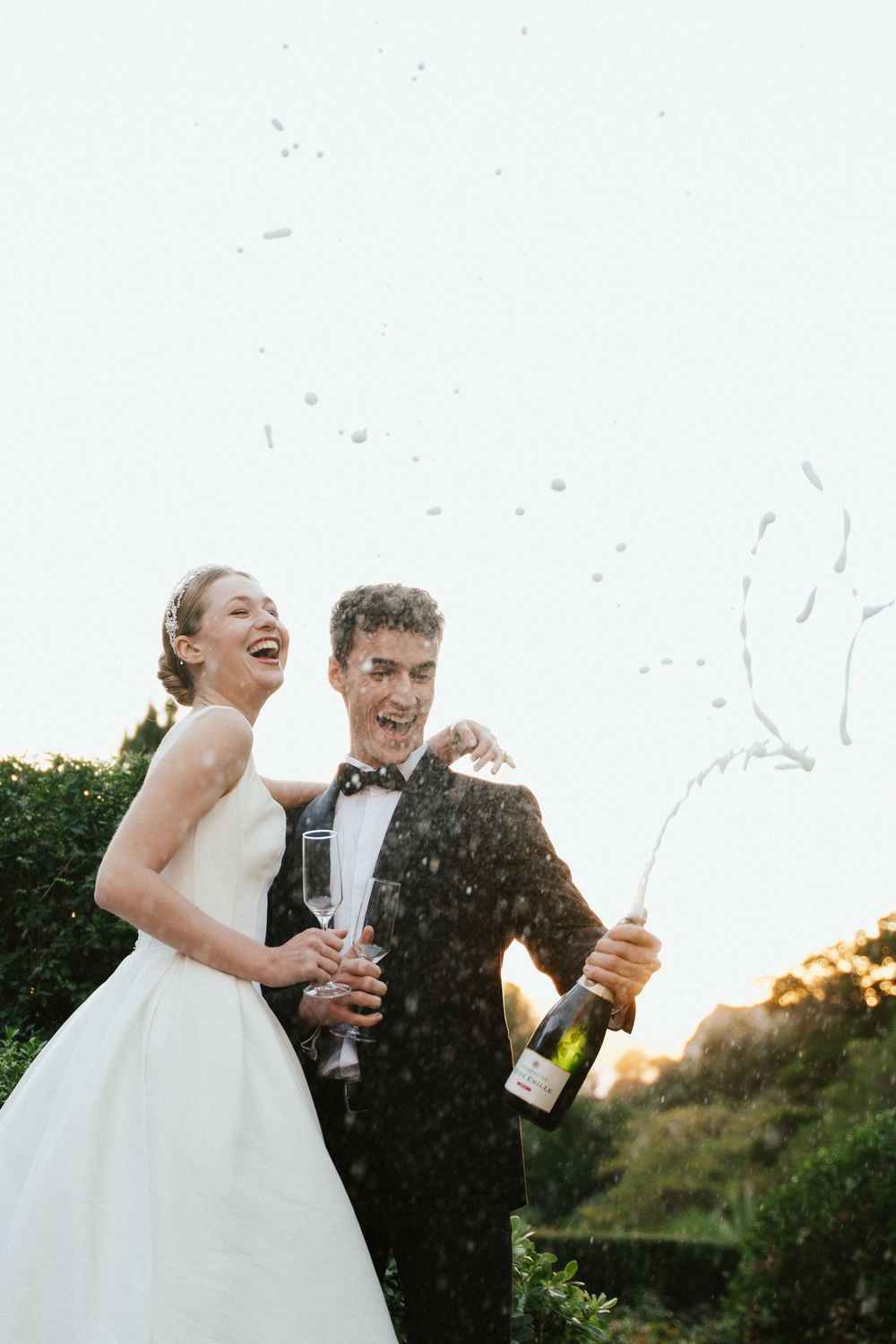 Joyful bride and groom popping champagne bottle outdoors, celebrating their wedding day with laughter and bubbly spray.