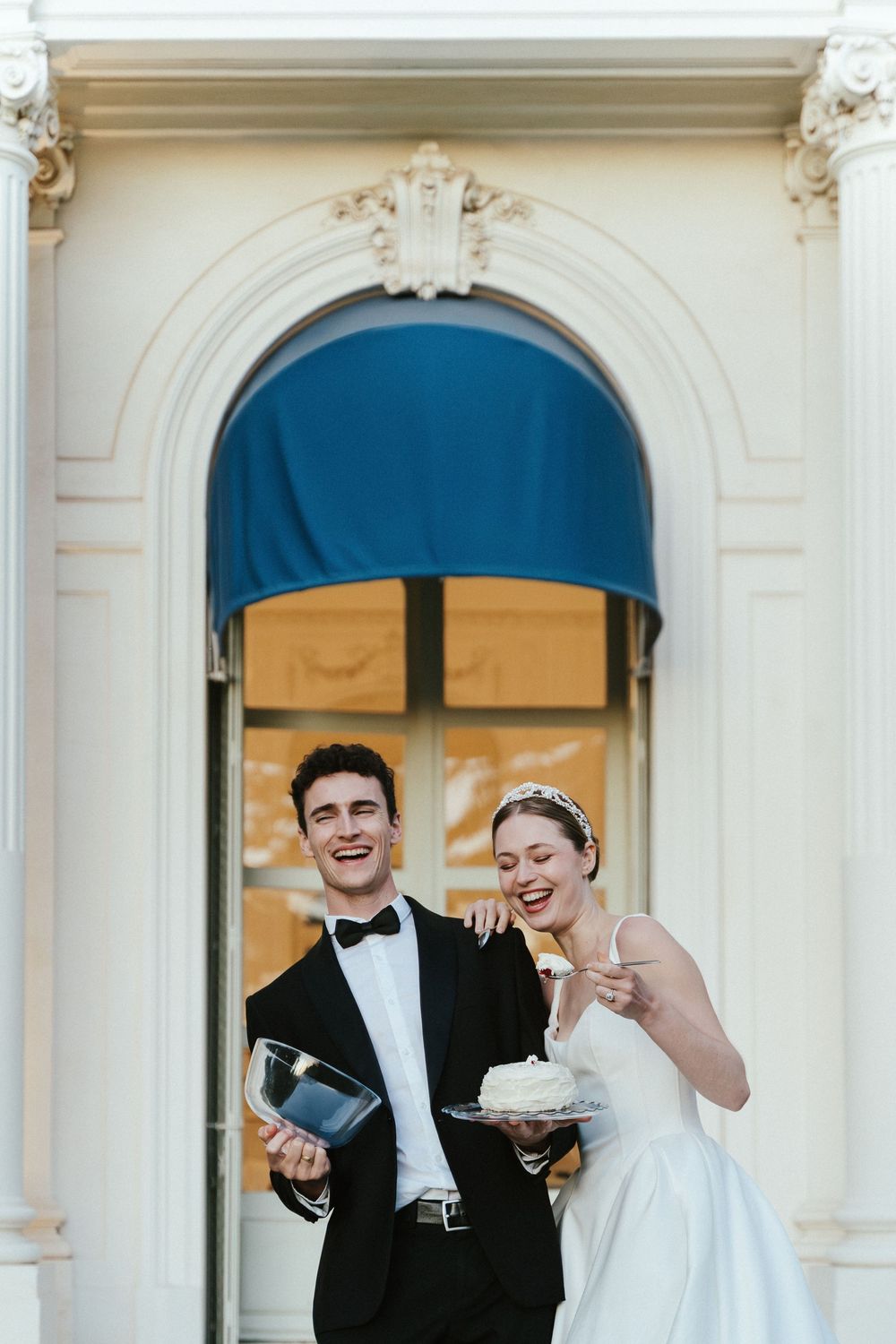 Smiling bride and groom cutting wedding cake outside elegant white building with blue awning.