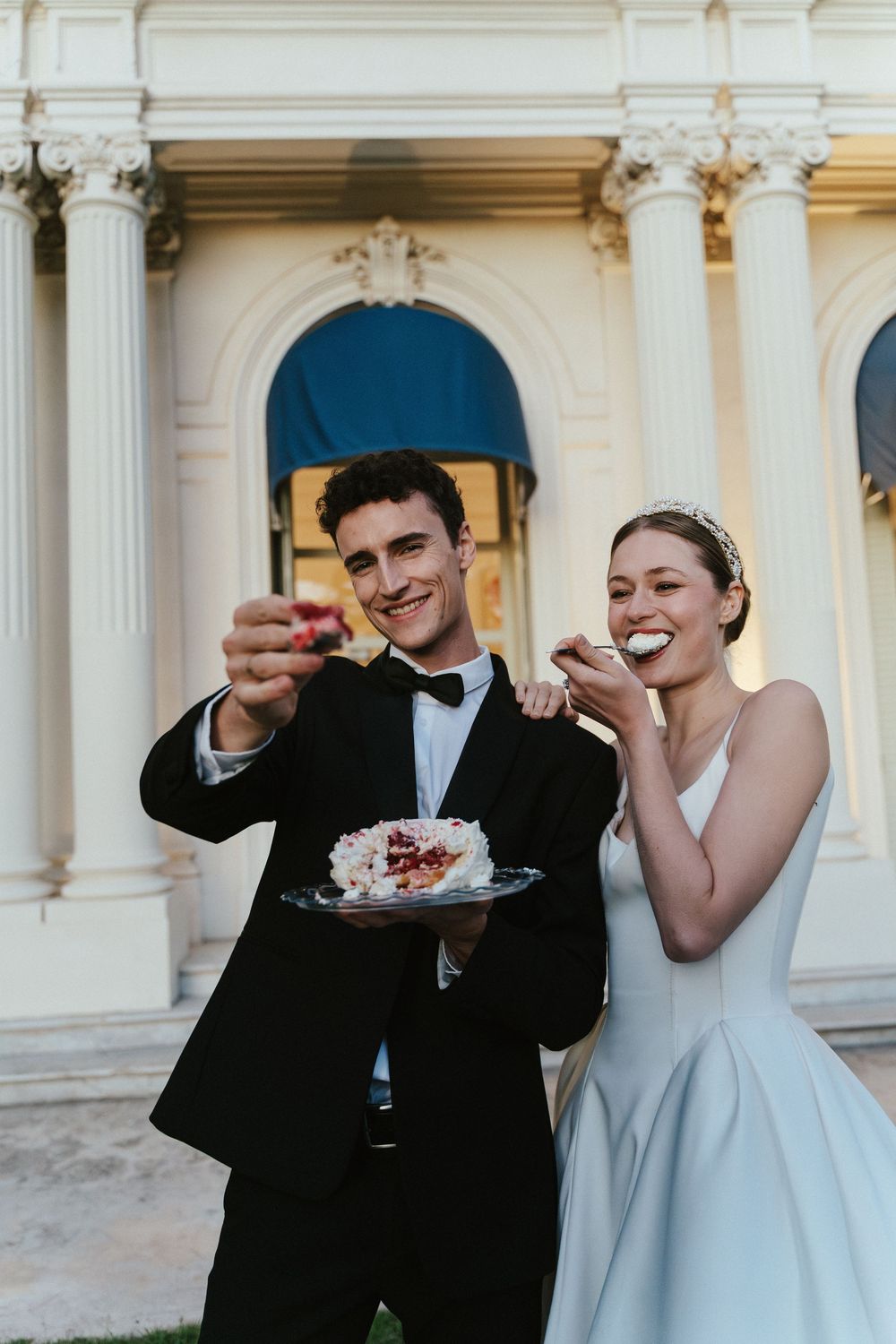 Bride and groom sharing wedding cake outside elegant white building with columns, smiling and enjoying their celebration.