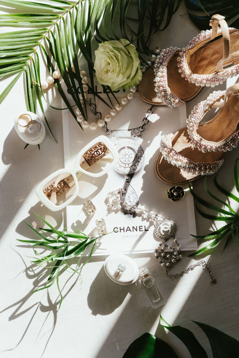 Chanel flatlay with pearl sandals, sunglasses, jewelry, white rose, and tropical leaves on white surface.