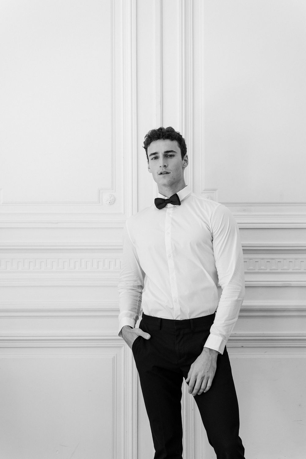 Young man in white dress shirt and black bow tie posing confidently against ornate white wall paneling.