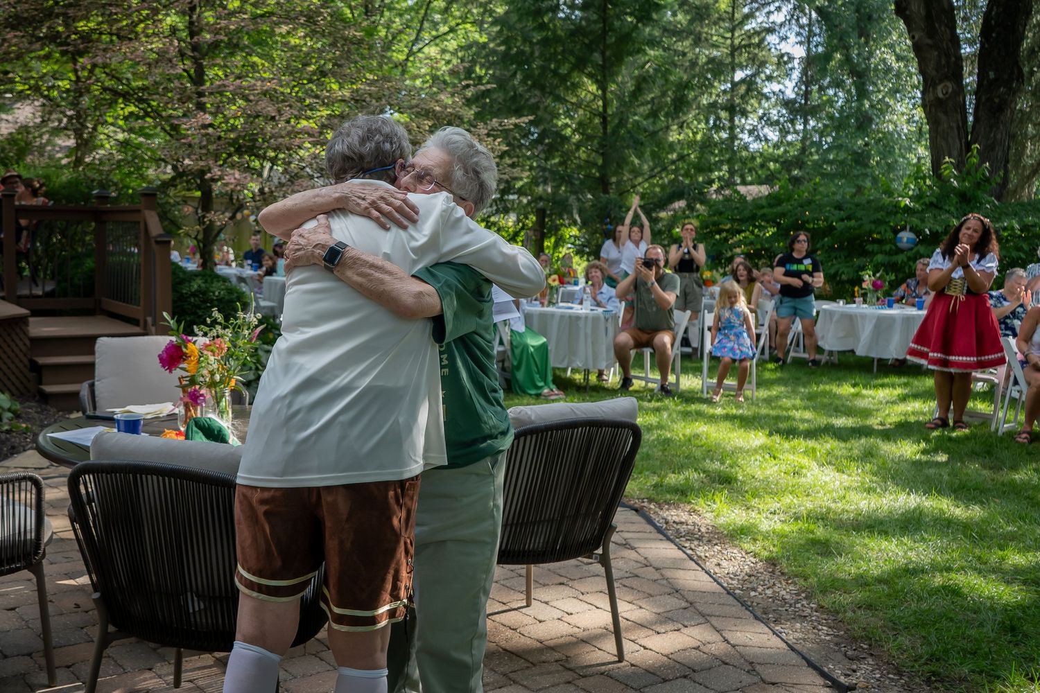 People embrace during an outdoor gathering in a lush garden setting with festive decorations and attendees.