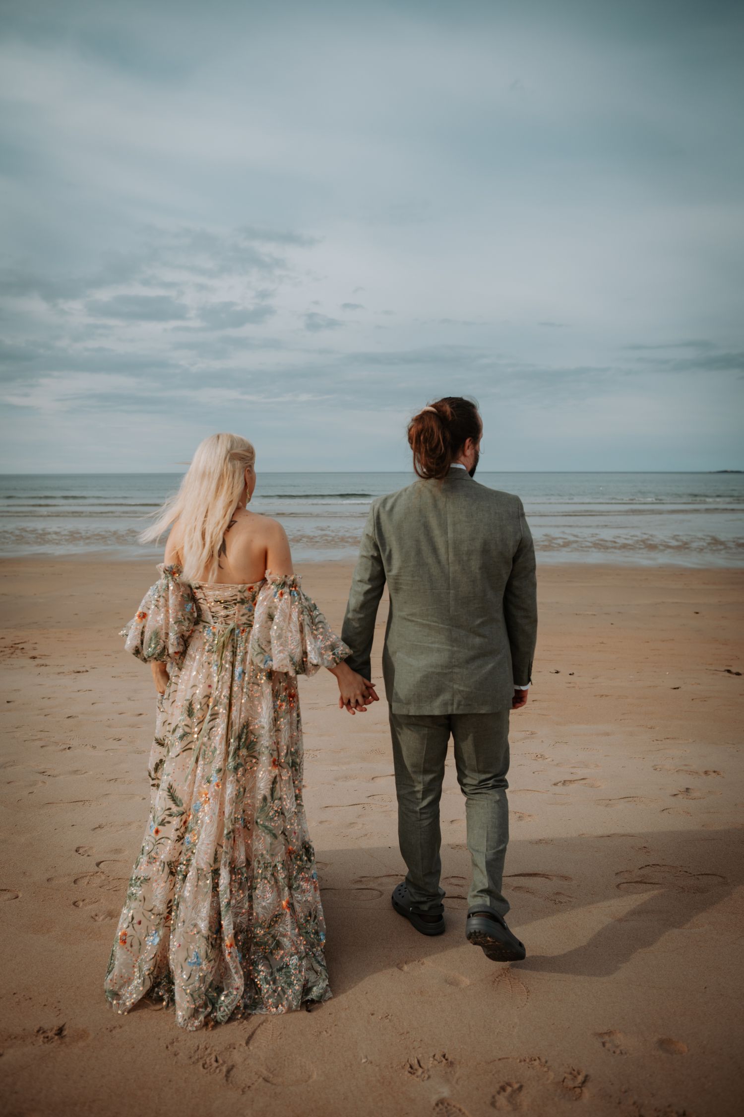 A couple holding hands looks out at the ocean on a cloudy day, standing on a sandy beach in casual attire.