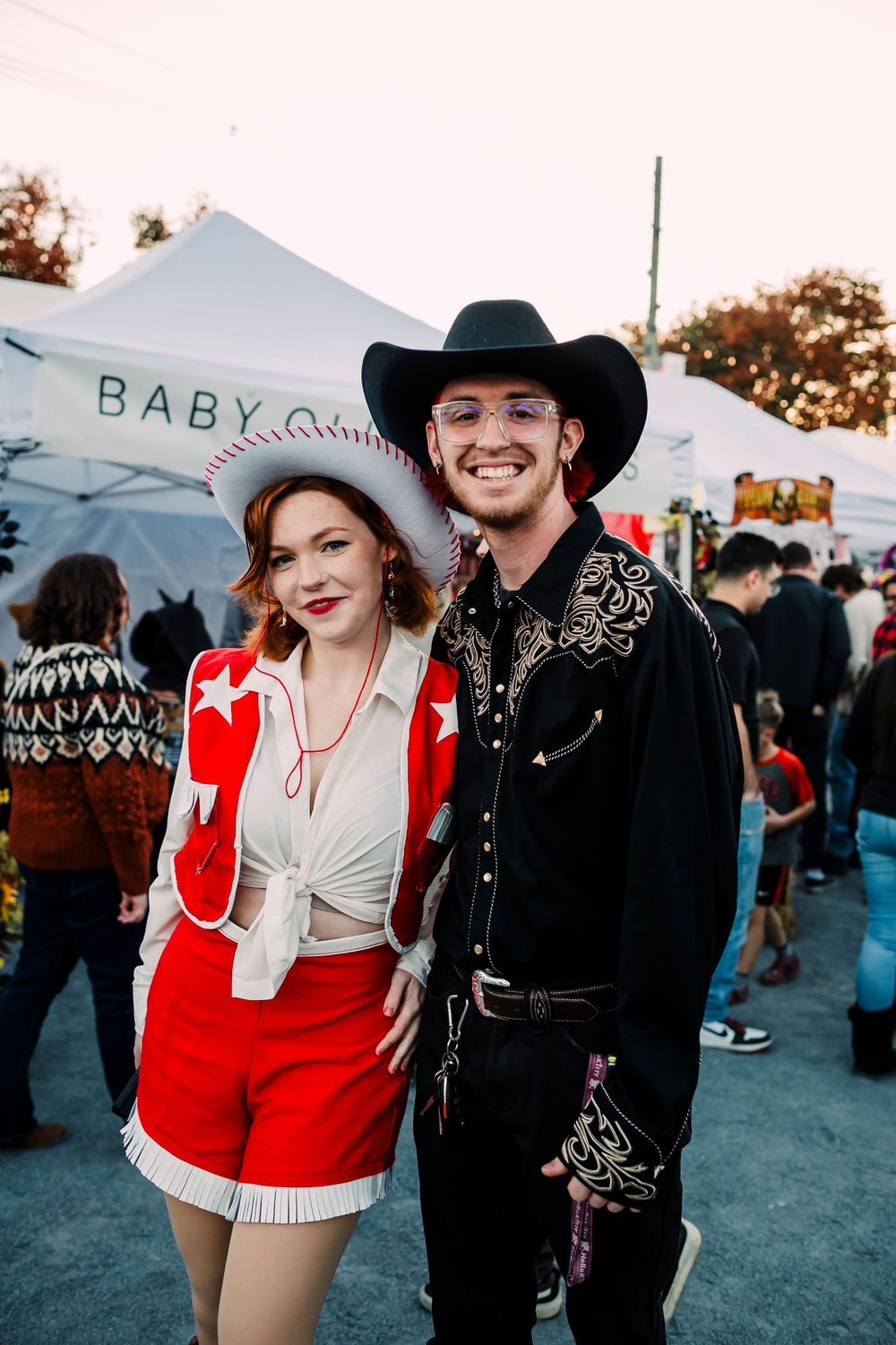 A couple in western attire stands at an outdoor festival, with her in red and him in a black embroidered outfit and cowboy hat.