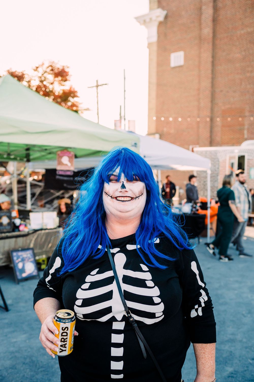 Person in Halloween skeleton costume with bright blue hair holding a drink can at an outdoor festival.