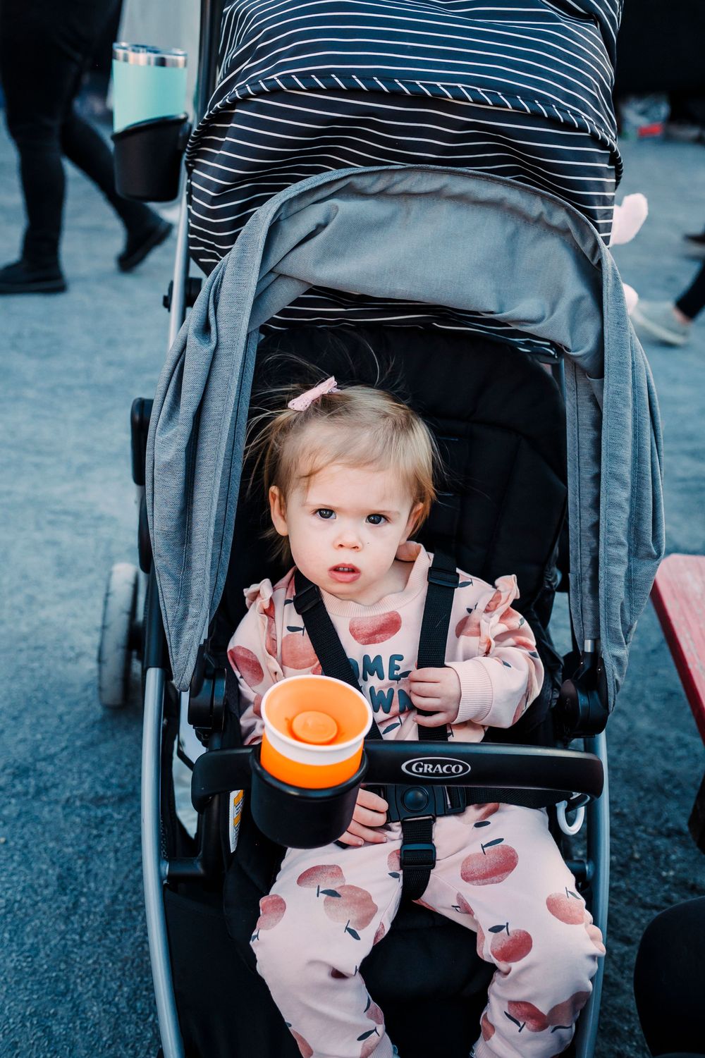 Baby sits in a black stroller holding an orange snack cup during an outdoor event.