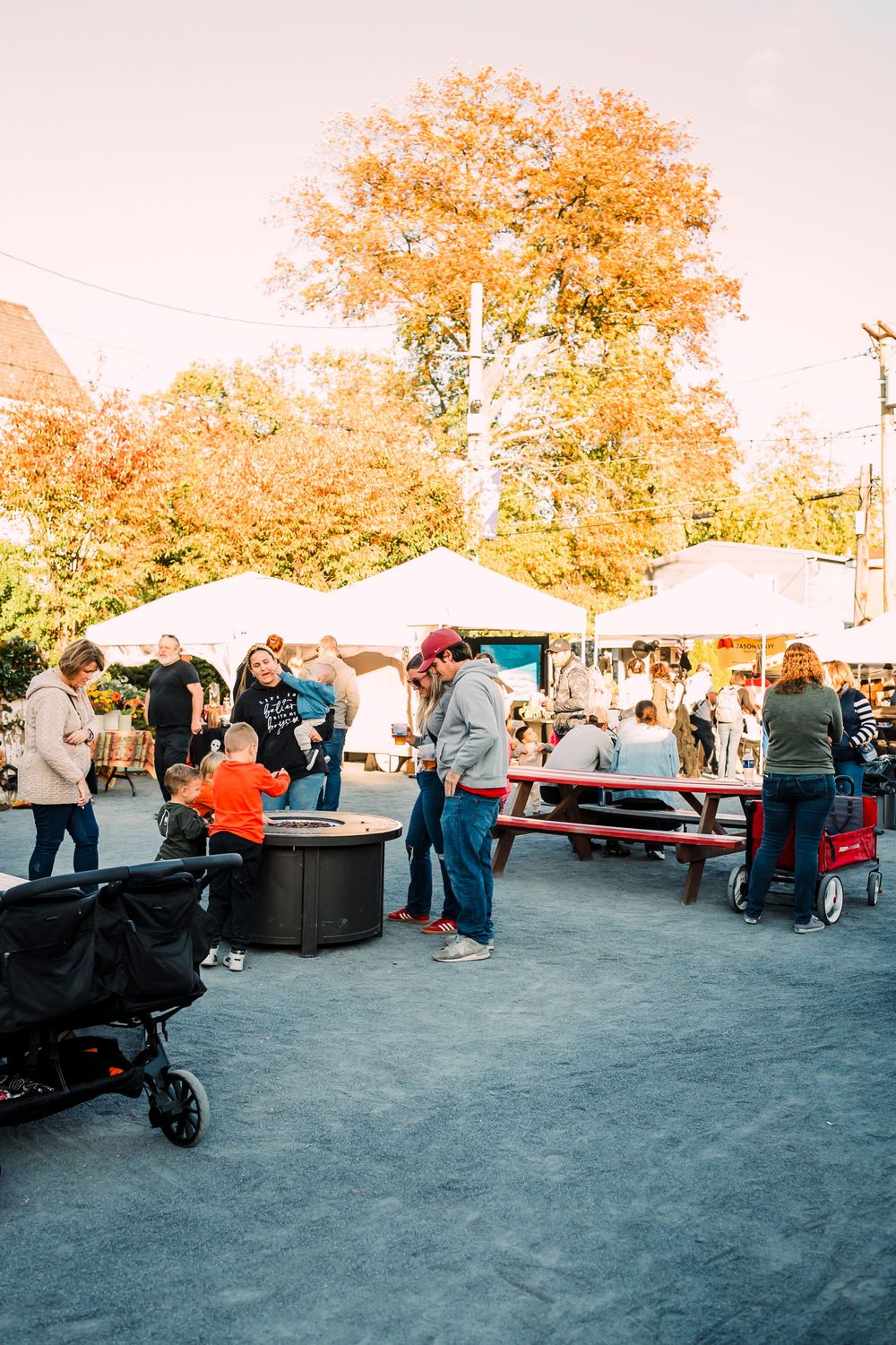 People browsing outdoor market stalls with autumn trees in the background.