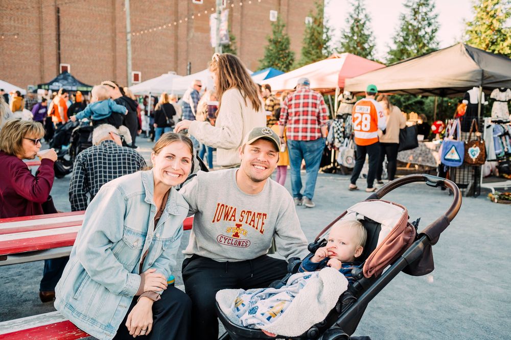 Family enjoying time together at an outdoor market with vendor tents and crowds visible in the background.