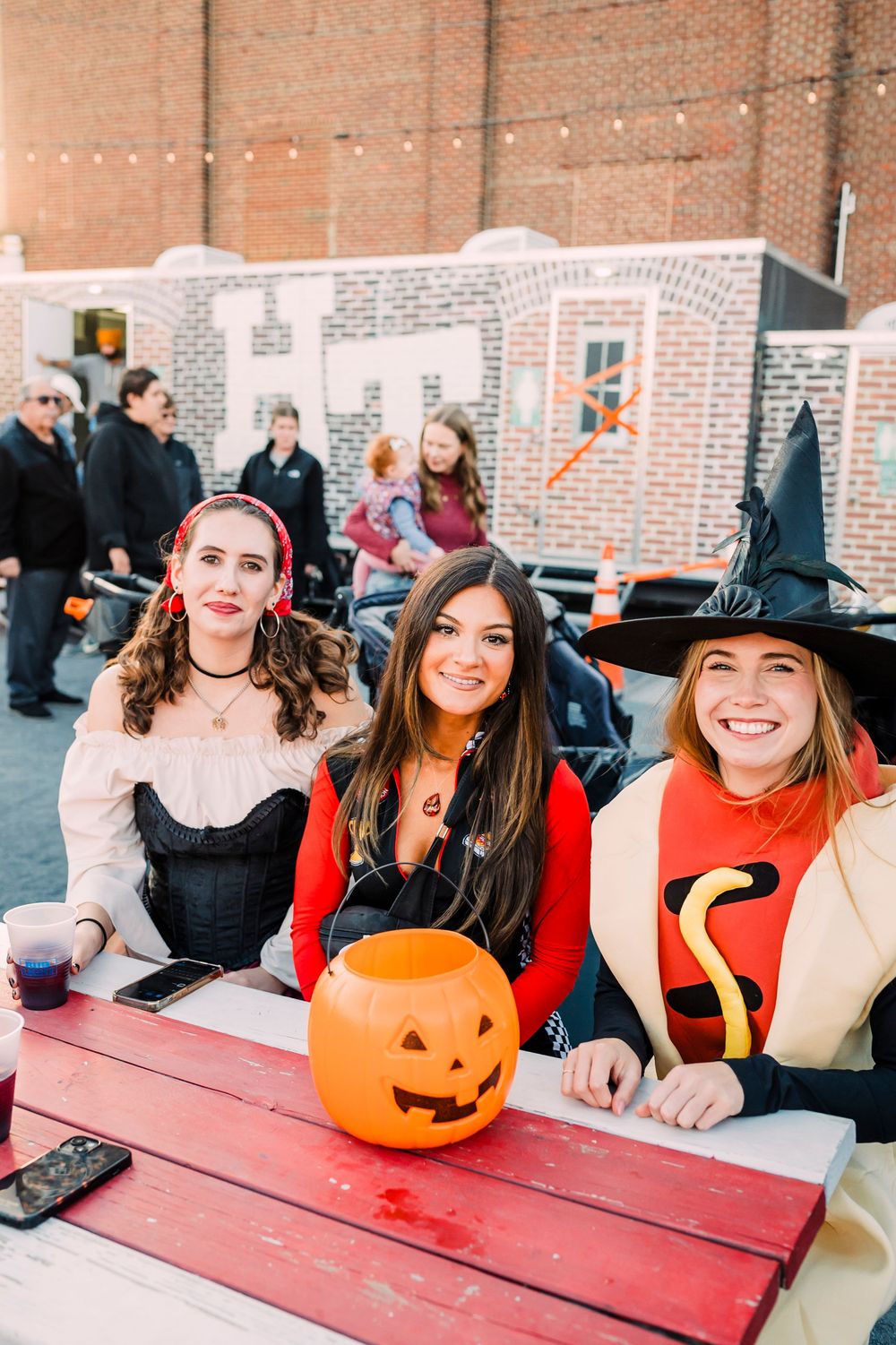 Group in Halloween costumes sitting at a picnic table with a jack-o'-lantern at an outdoor festival.