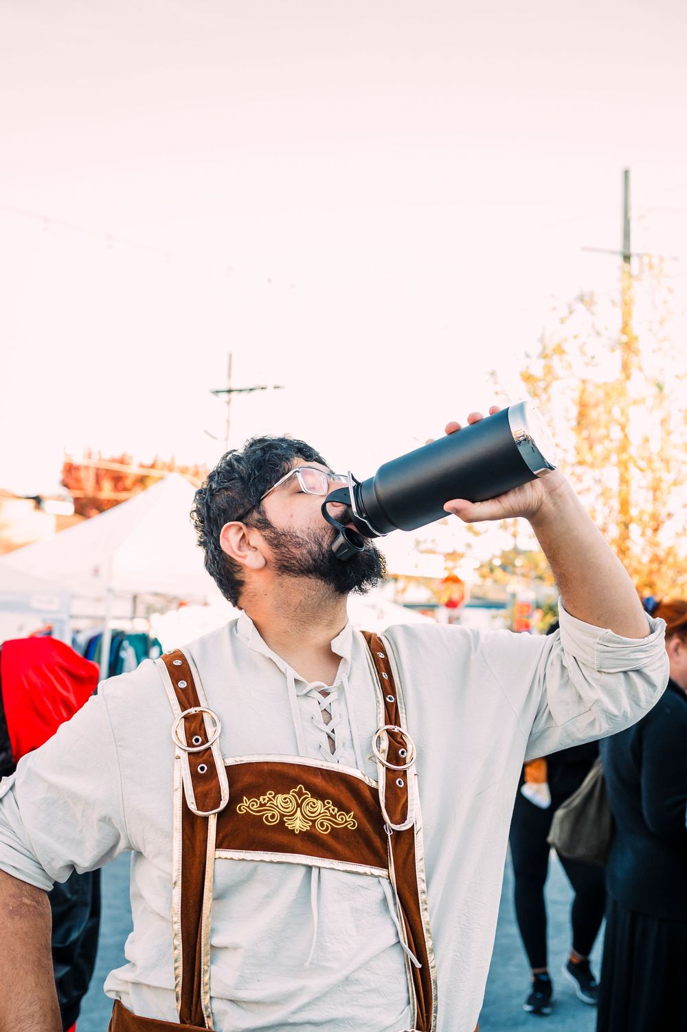 Person in traditional German lederhosen drinking from a large bottle at a festival.