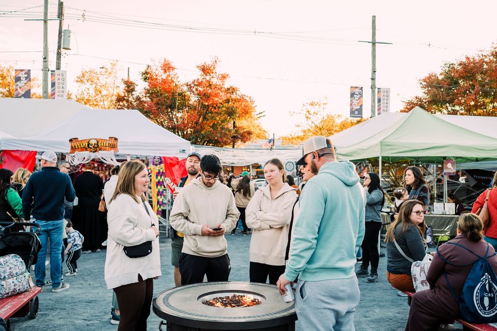 Crowd of people enjoying an outdoor event with white tents and fall foliage.