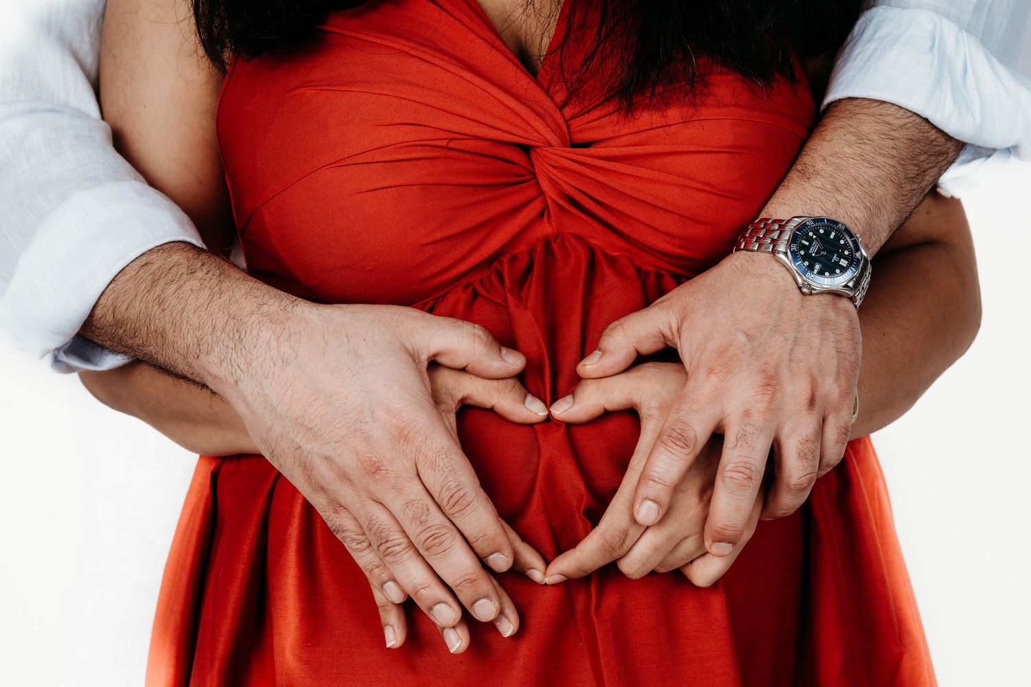 Hands form a heart shape against a bright red dress, with one person wearing a watch.