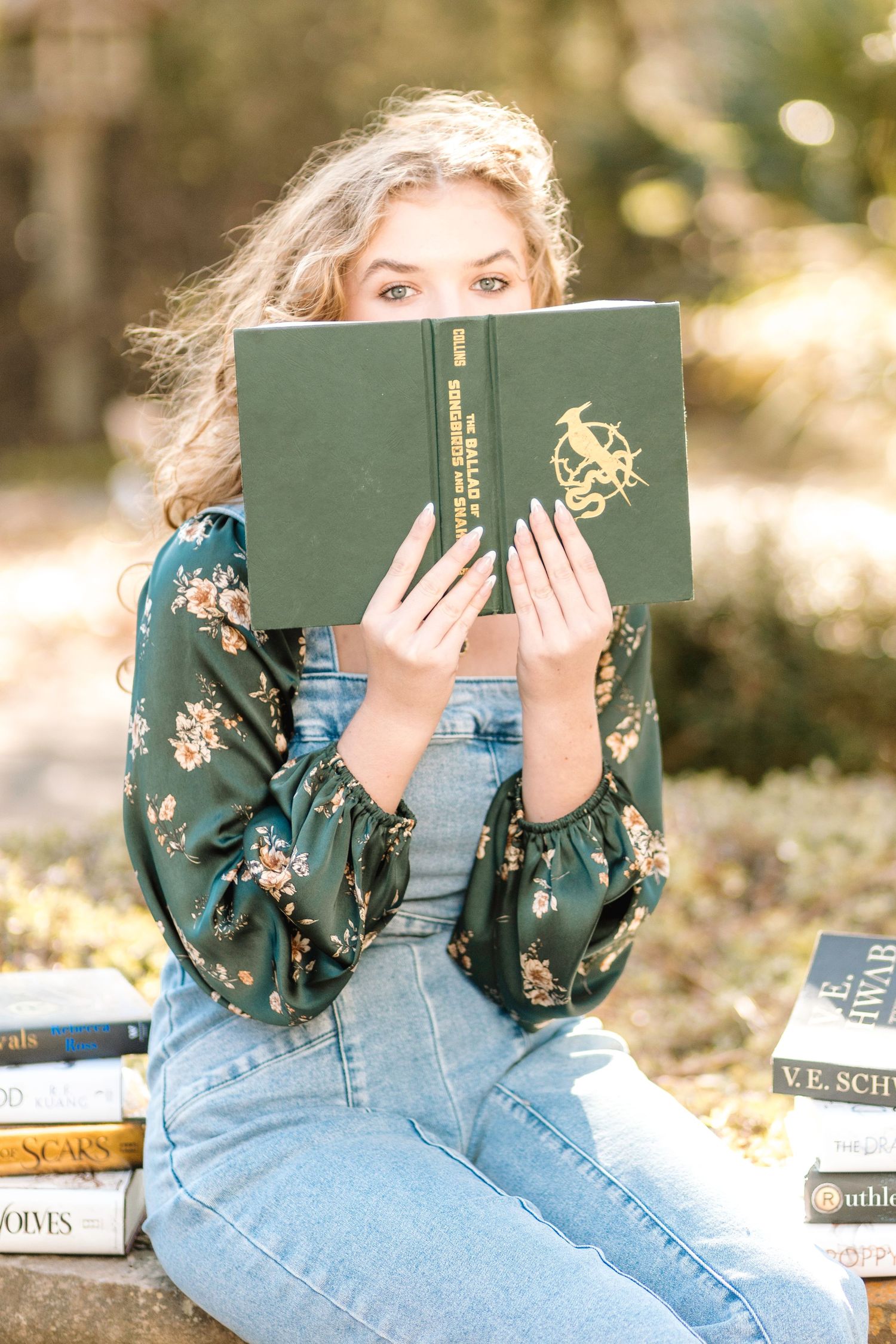 Reader in denim overalls and floral print shirt holds green hardcover book with golden emblem outdoors on sunny day.