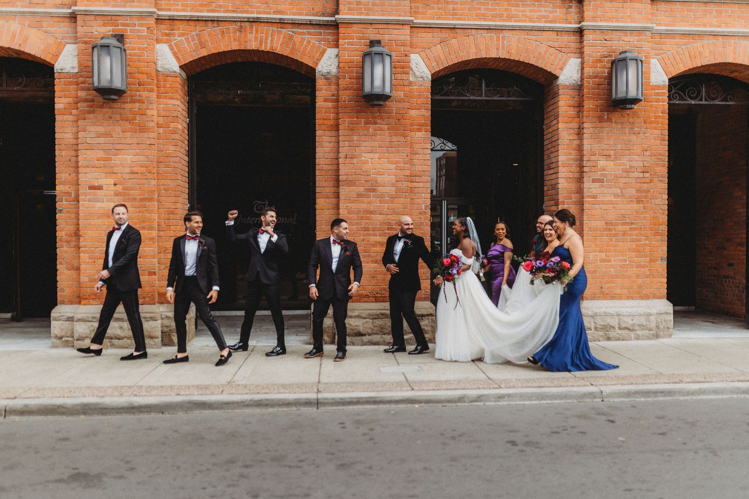 A wedding party poses along a rustic brick building with arched doorways and vintage wall sconces.