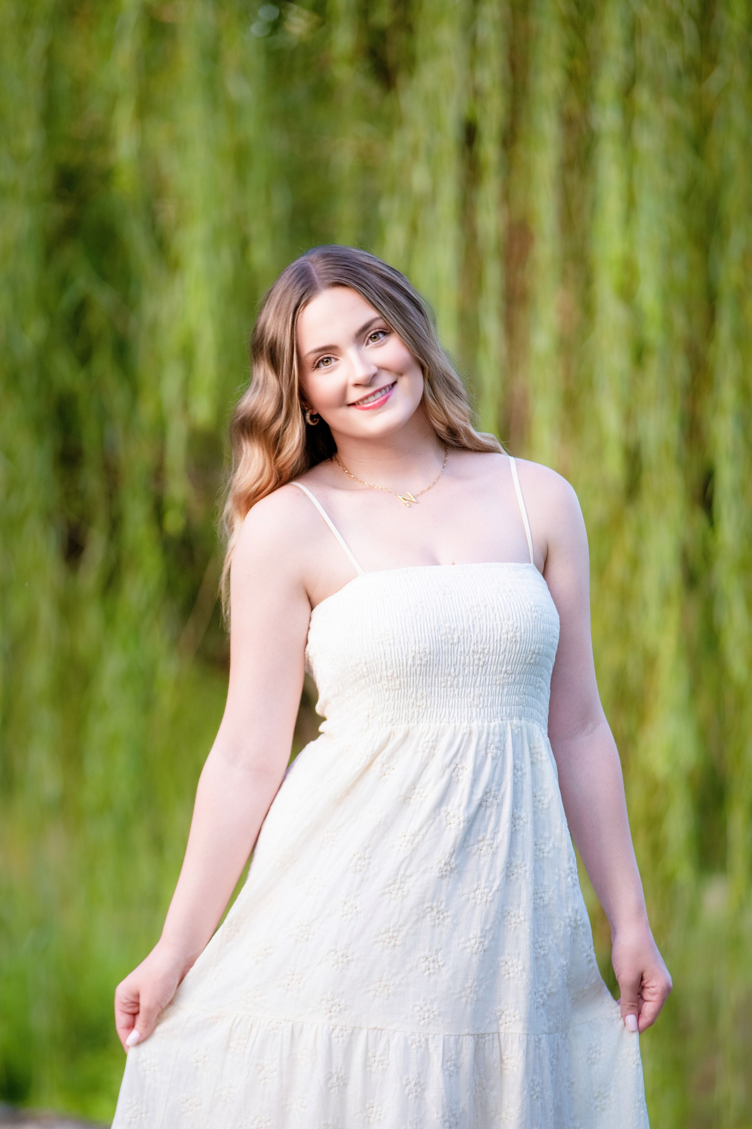 A white sundress with spaghetti straps is shown outdoors against a soft green willow tree background.