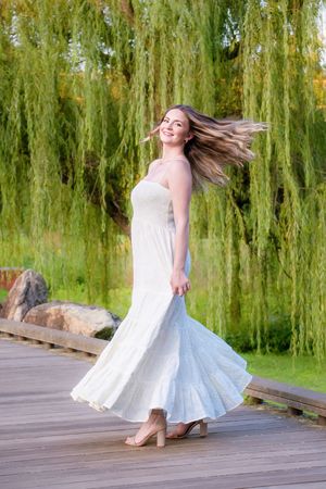 A white sundress twirls gracefully on a wooden dock against a backdrop of cascading willow trees.