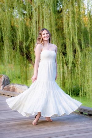 Someone in a flowing white summer dress twirls gracefully on a wooden dock with willow trees in the background.
