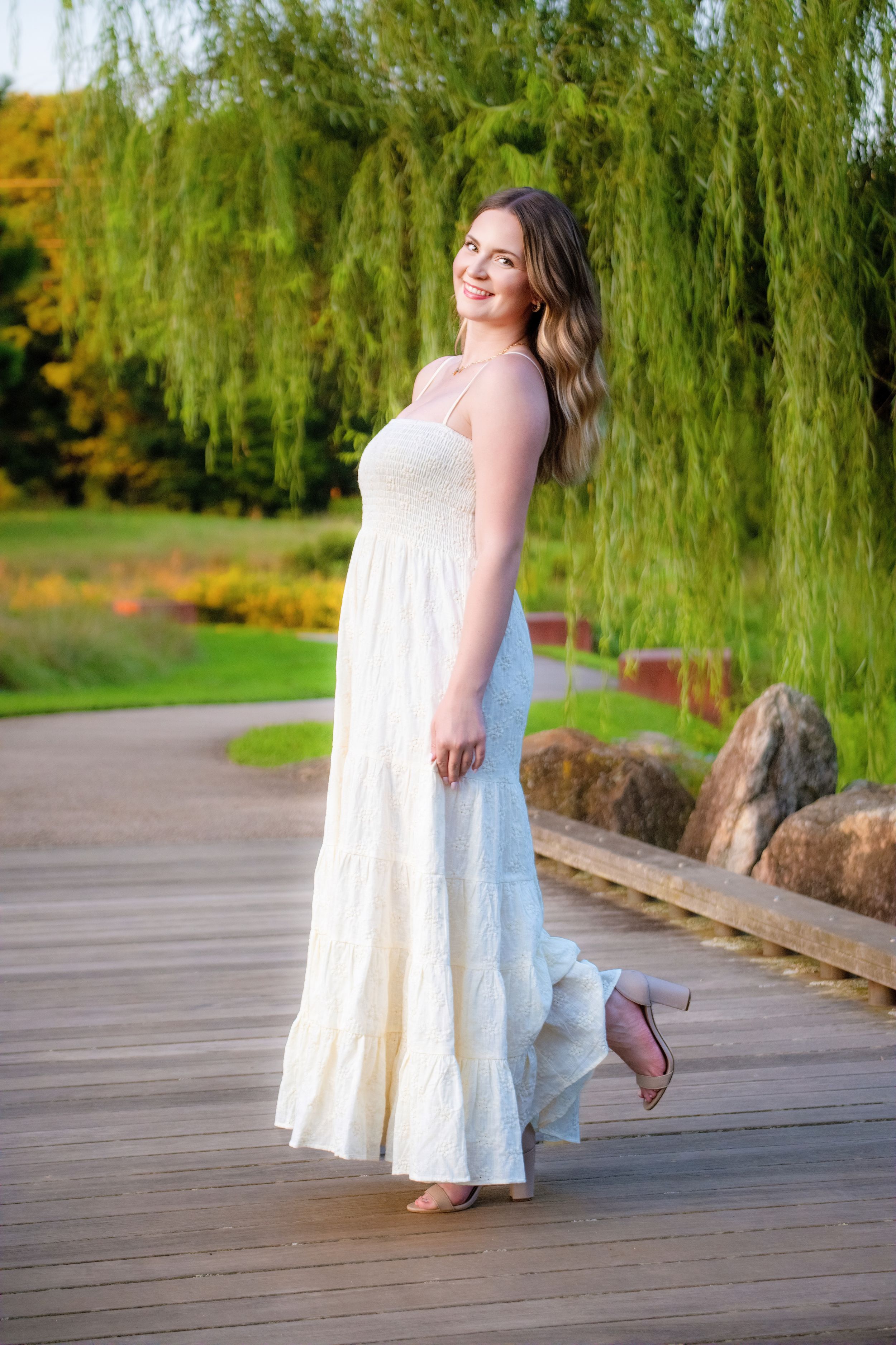 A flowing white maxi dress stands out against a backdrop of weeping willow trees and a wooden boardwalk in a garden setting.