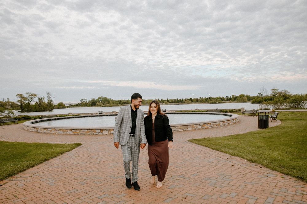 Two people walk along a curved brick pathway next to a waterfront park on a cloudy day.