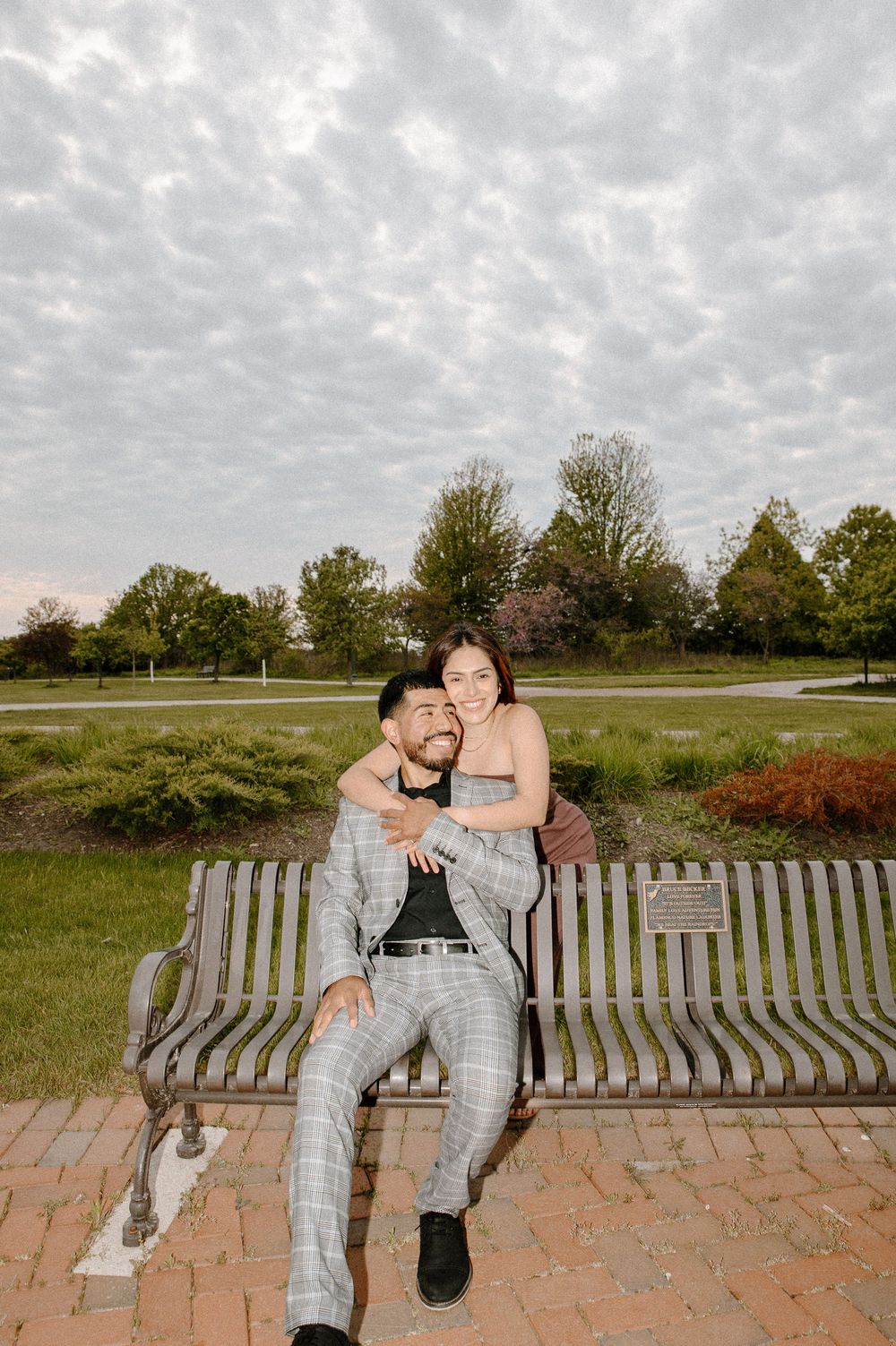 A couple shares an intimate moment sitting together on a wooden bench in an outdoor park setting with cloudy skies.