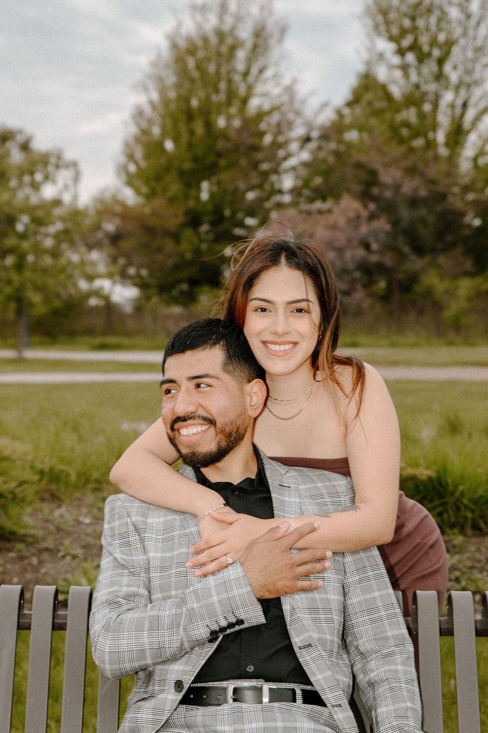 A couple laughs and embraces while sitting on a wooden bench in a park setting.