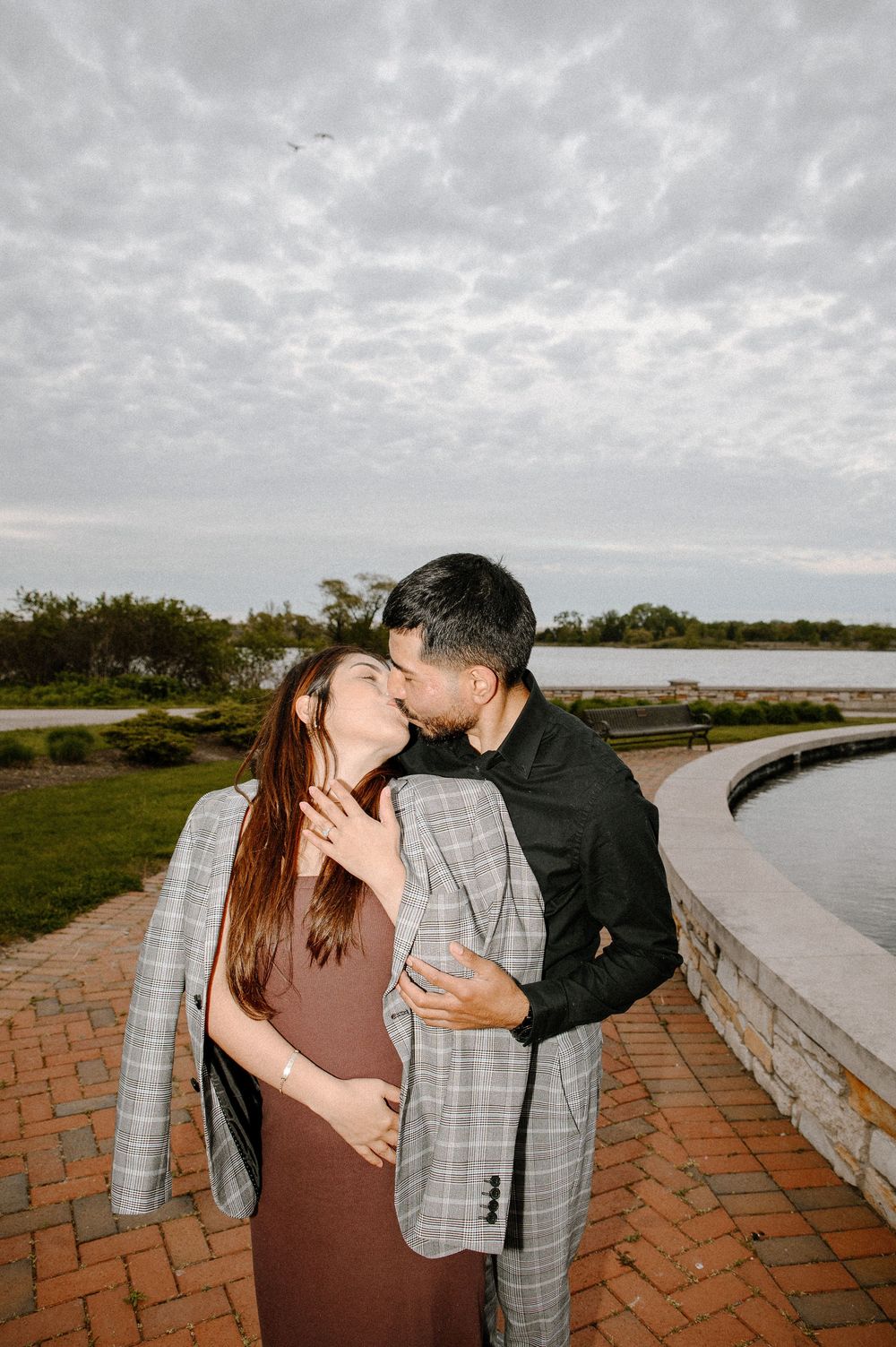 Two silhouettes share an intimate moment on a brick path against a cloudy sky near a waterway.