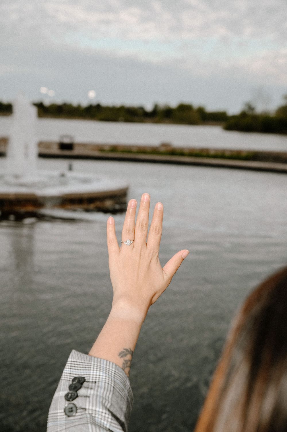 A hand with a new engagement ring is held up against a waterfront backdrop.