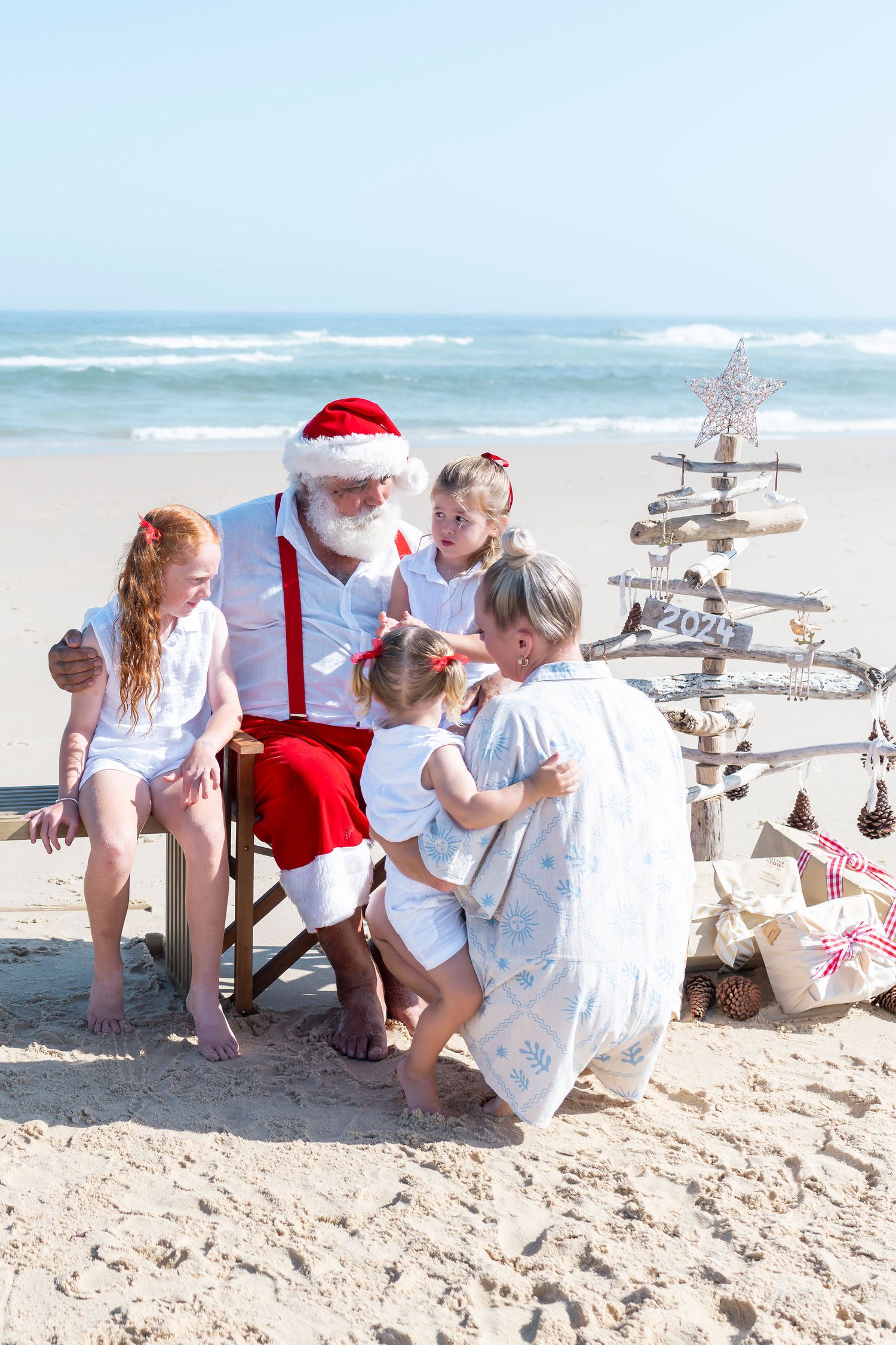 A child wearing a Santa hat and light overalls points while standing on a sunny beach during the holiday season.
