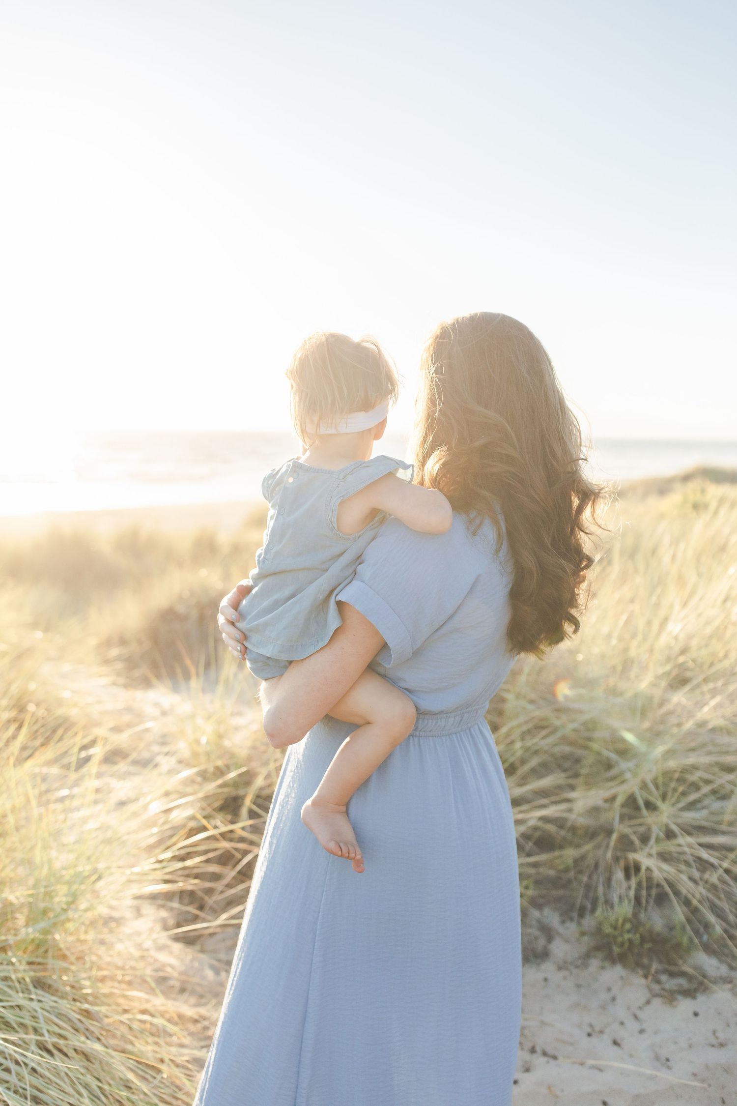 A figure in a flowing blue dress holding a small child gazes at a sunset on the sandy beach.Film by Chelsee Rawe Photography