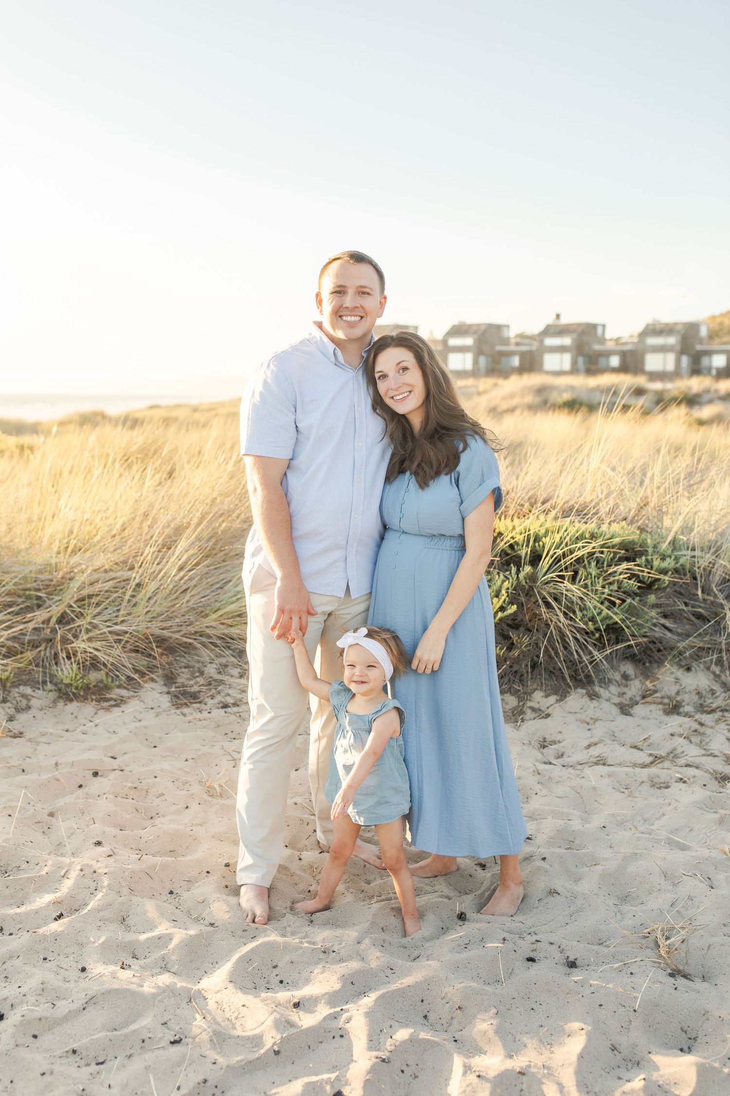 A family shares tender moments on a sandy beach at sunset with coastal houses in the background.Film by Chelsee Rawe Photography