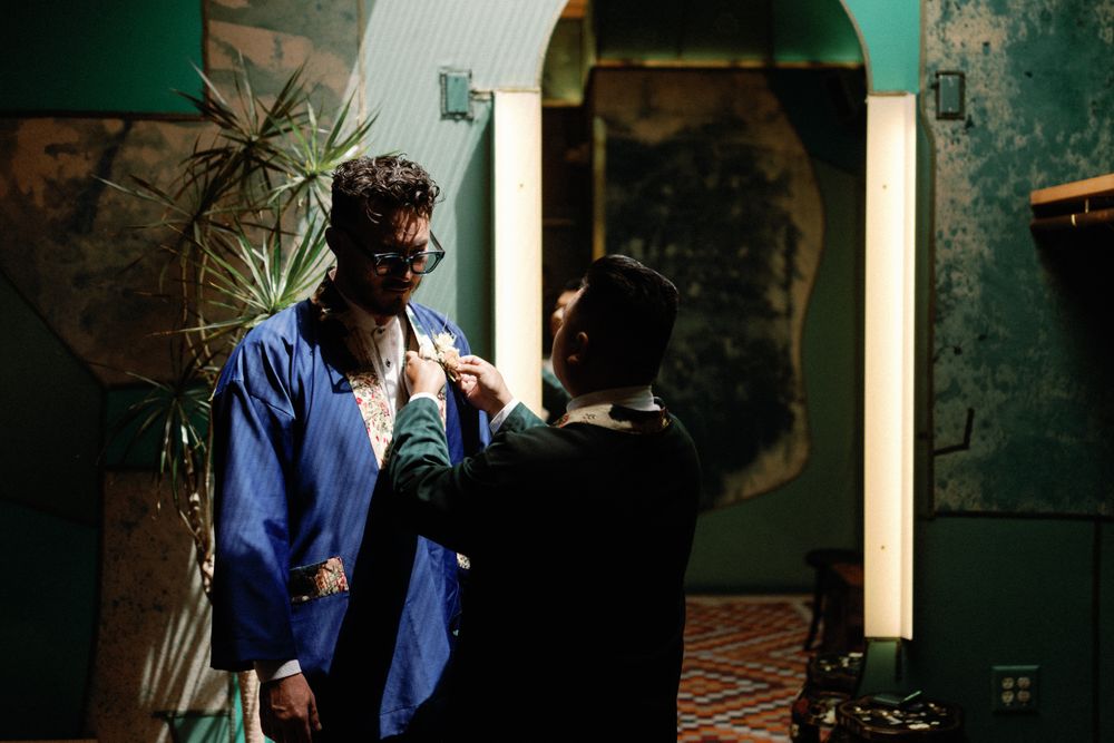 Groom in blue robe gets boutonniere pinned by groomsman in dramatic moody green venue before wedding.