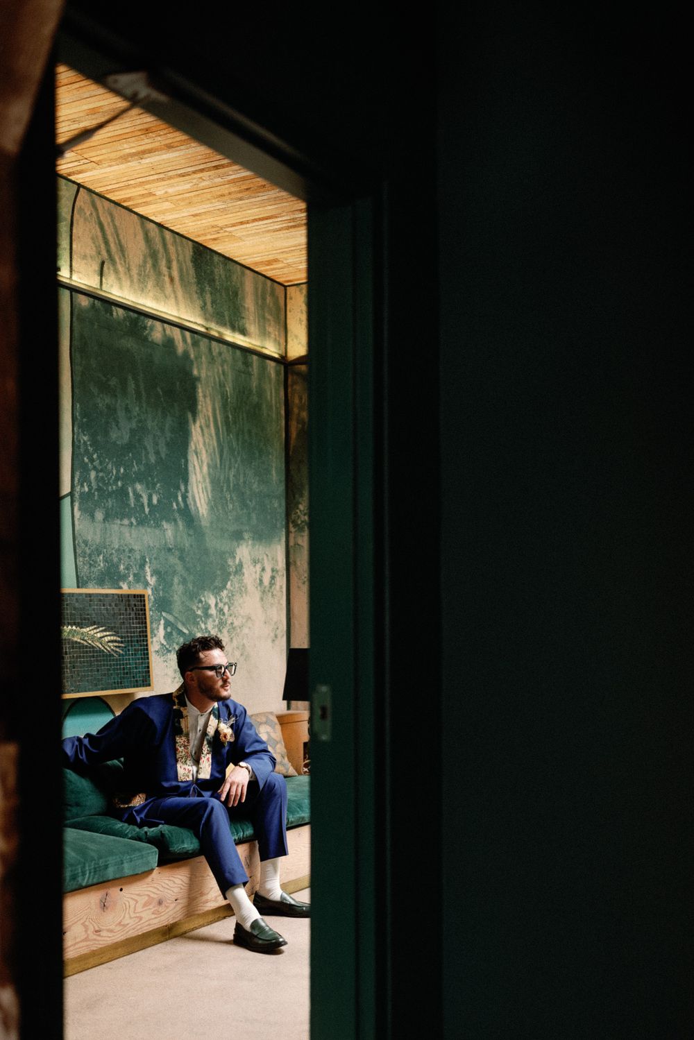 Man in blue suit sitting on green velvet sofa viewed through doorway in stylish moody interior.