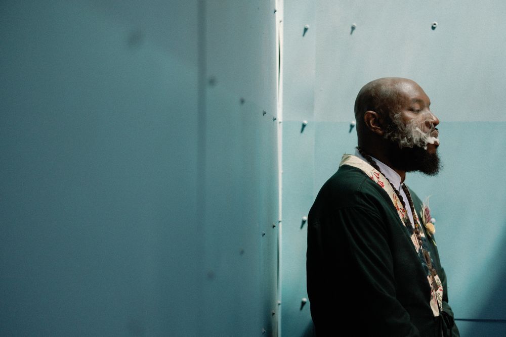 Side profile of bearded Black man in dark jacket standing against light blue wall outdoors.