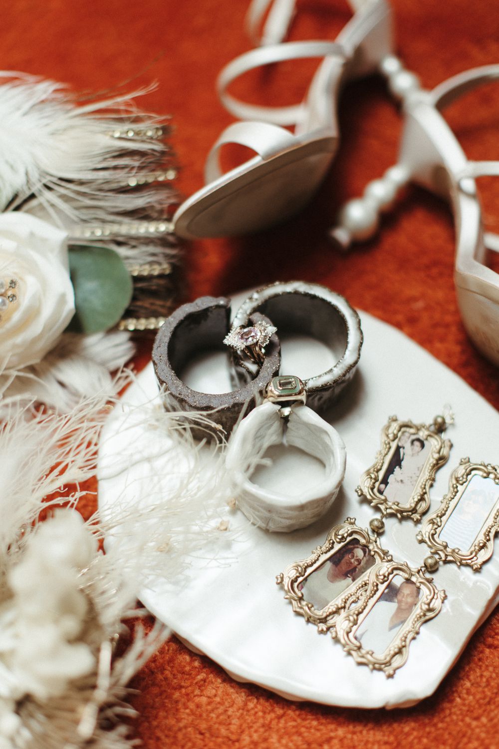 Wedding rings and jewelry displayed on white dish with feathers and bridal heels on orange surface.