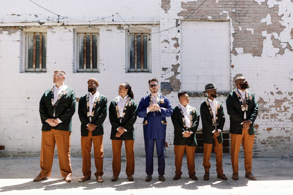 Groom in blue suit stands with groomsmen in matching black jackets and brown pants outdoors.